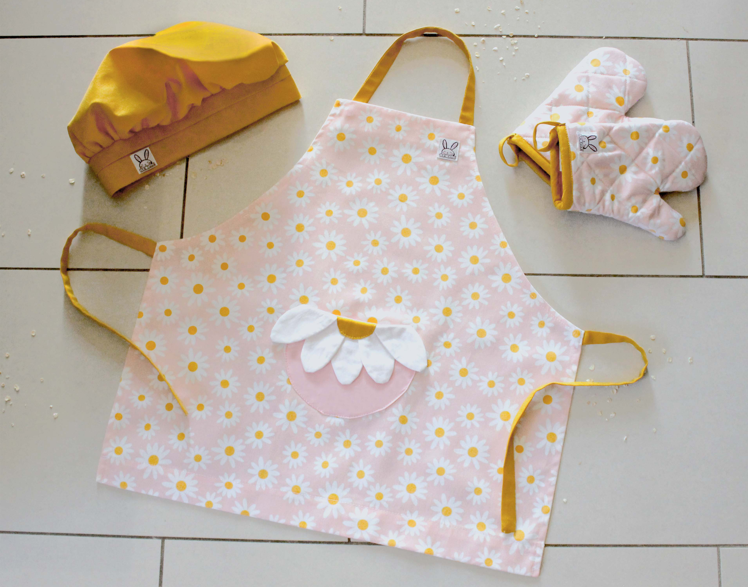 Ensemble de ''mini-chef'' pour faire fureur dans la cuisine ! Le tout comprend: 1 tablier, 1 paire de mitaine et 1 chapeau ajustable. Le tablier fleuri s'ajuste à l'encolure avec des boutons pressions. Une poche amusante en forme de fleur au devant. Produits fait au Québec. Les mitaines de four sont isolées. Il y a des ganses pour les accrocher sur un crochet. La toque jaune curry est ajustable à l'aide d'un élastique.