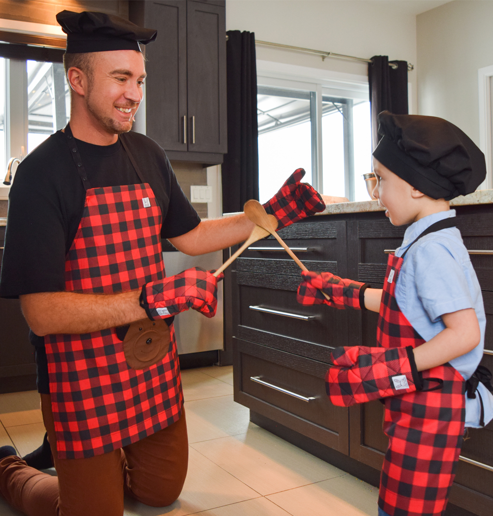 Un garçon et son papa s'amuse à cuisiner ensemble. Ils portent, tous les 2, l'ensemble de tablier bûcheron rouge et noir. Des mitaines de four qui s'agence avec le tablier et le chapeau de chef noir pour finaliser le look d'un vrai cuisinier.