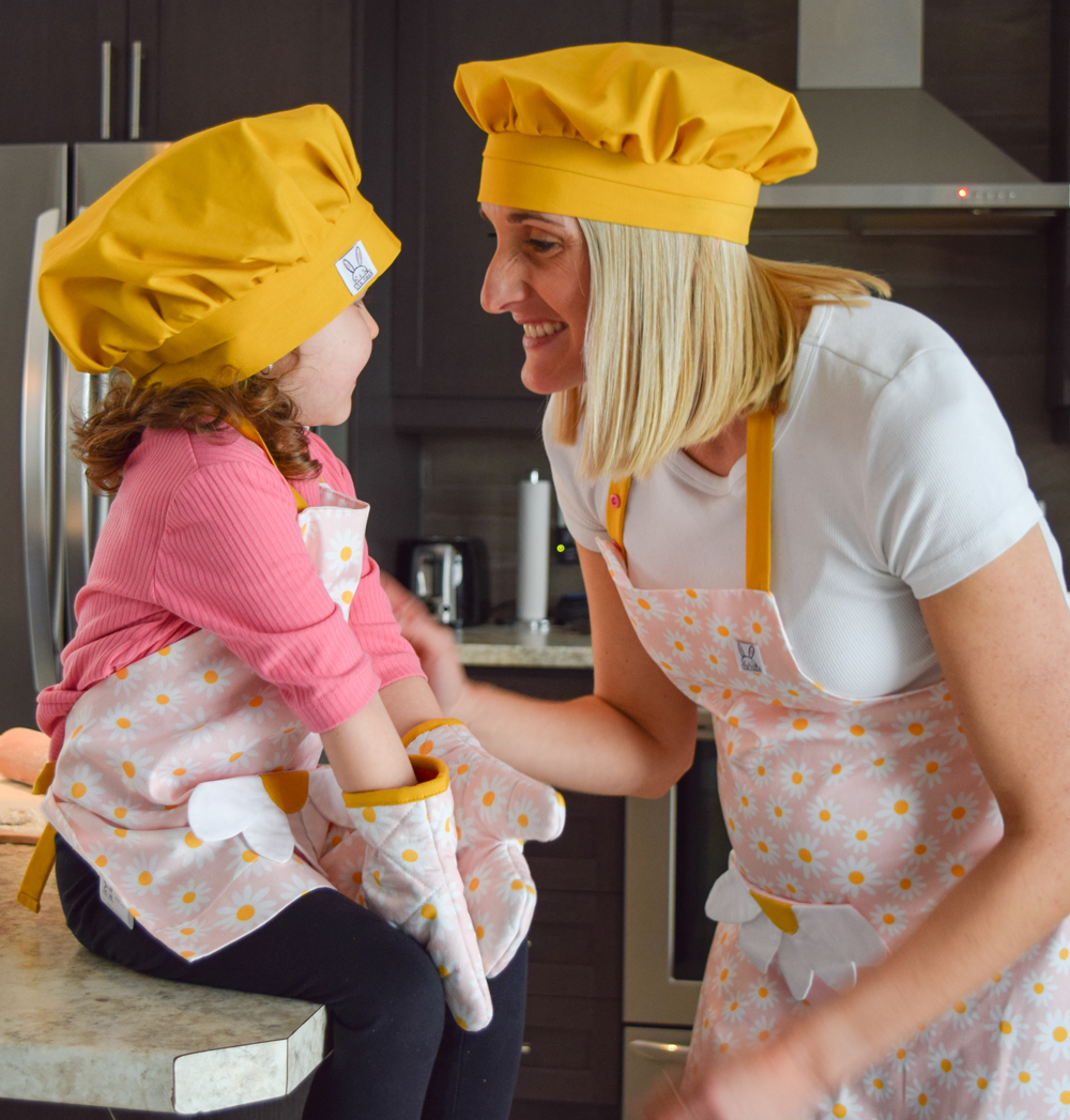 Une maman avec sa fille fait la cuisine ensemble. Elles portent le même ensemble de cuisine fleuri rose et jaune. Une poche amusante au devant en forme de fleur. Des mitaines de four isolés et un chapeau de chef jaune curry pour finaliser le look d'un vrai cuisinier.