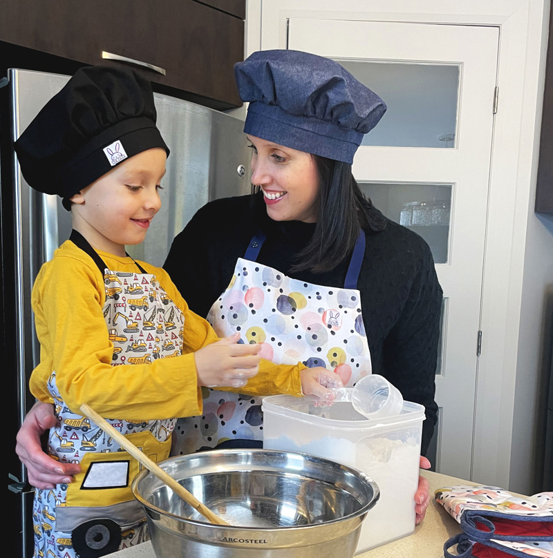 Une petit garçon et une maman cuisine ensemble. Le petit garçon porte un tablier de camion avec un chapeau de chef noir et la maman porte le tablier à pois avec une toque en denim.
