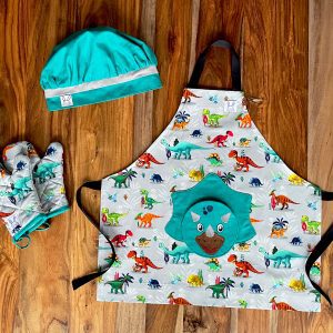 Ensemble de tablier à motif de dinosaures. Le tablier s'ajuste à l'encolure avec des boutons pressions. Une poche amusante en forme d'un tricératops au devant. Les mitaines de four sont isolées. Il y a des ganses pour les accrocher sur un crochet. La toque de couleur émeraude est ajustable à l'aide d'un élastique. Produits fait au Québec