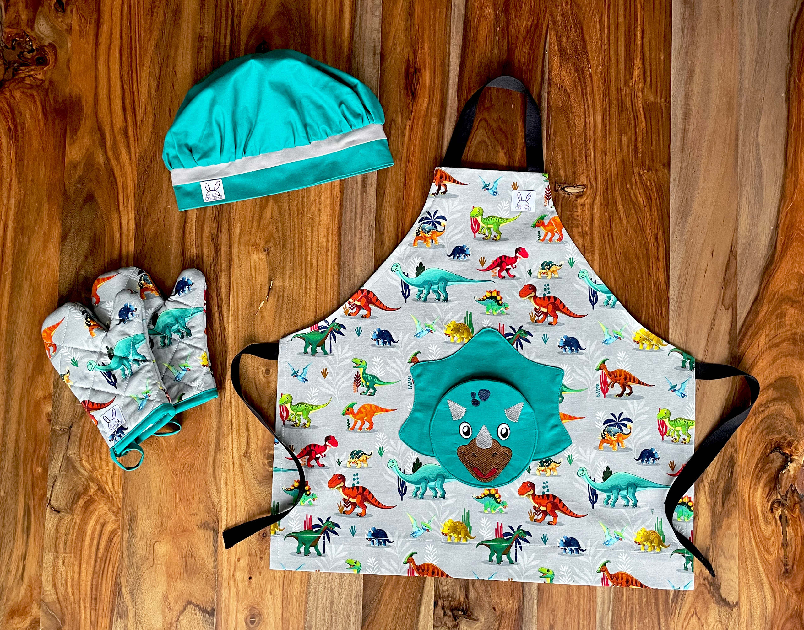 Ensemble de tablier à motif de dinosaures. Le tablier s'ajuste à l'encolure avec des boutons pressions. Une poche amusante en forme d'un tricératops au devant. Les mitaines de four sont isolées. Il y a des ganses pour les accrocher sur un crochet. La toque de couleur émeraude est ajustable à l'aide d'un élastique. Produits fait au Québec