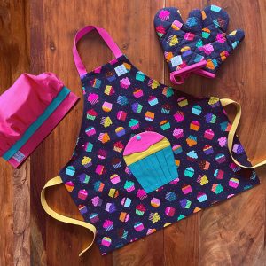 Ensemble de tablier à motif de petits gâteaux. Le tablier s'ajuste à l'encolure avec des boutons pressions. Une poche amusante en forme d'un gros cupcakes au devant. Les mitaines de four sont isolées. Il y a des ganses pour les accrocher sur un crochet. La toque de couleur fuchsia est ajustable à l'aide d'un élastique. Produits fait au Québec