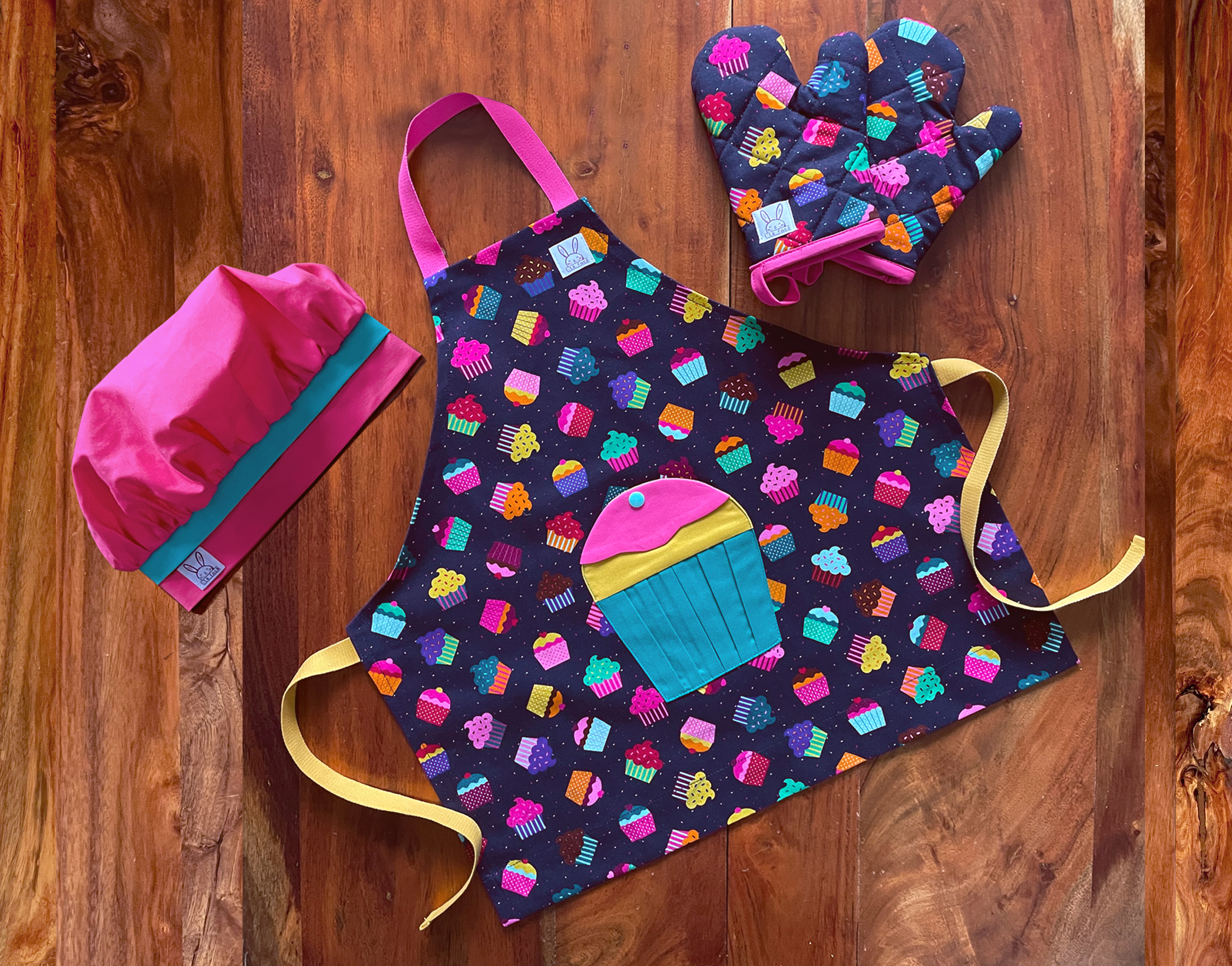 Ensemble de tablier à motif de petits gâteaux. Le tablier s'ajuste à l'encolure avec des boutons pressions. Une poche amusante en forme d'un gros cupcakes au devant. Les mitaines de four sont isolées. Il y a des ganses pour les accrocher sur un crochet. La toque de couleur fuchsia est ajustable à l'aide d'un élastique. Produits fait au Québec